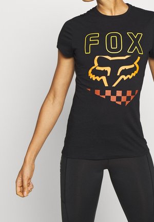 Personne portant un t-shirt noir ajusté avec un texte jaune "FOX", un logo tête de renard orange et un motif à carreaux orange et marron.