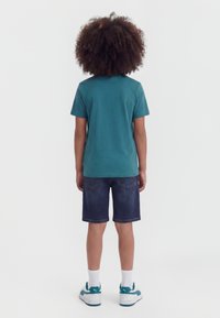 Παιδί με σγουρά μαλλιά στέκεται με την πλάτη γυρισμένη, φορώντας γαλάζιο κοντομάνικο μπλουζάκι, σκούρο τζιν σορτς, λευκές κάλτσες και γαλάζια-λευκά αθλητικά παπούτσια.