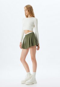 Jupe plissée vert olive avec une ceinture blanche, associée à un crop top blanc côtelé. Le mannequin porte des bottes à plateforme blanches épaisses.