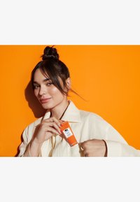 En oransje tube med hudpleieprodukt med en hvit etikett holdes av en person i en lys skjorte, mot en oransje bakgrunn.