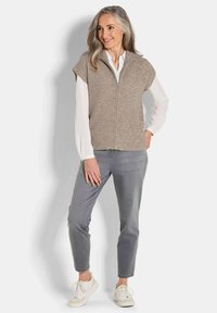 Beige Strickweste mit Reißverschluss, kurzen Puffärmeln, getragen über einem weißen Hemd und kombiniert mit grauen Hosen und weißen Sneakers.