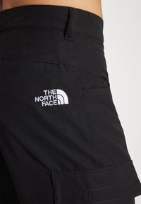 Čierne cargo nohavice s viditeľným bielym logom "The North Face", s hladkou textúrou a štruktúrovaným dizajnom, prezentujúce vrecká a švy.
