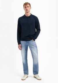 Marineblauwe trui met ritssluiting en lange raglanmouwen, gecombineerd met lichtblauwe denim jeans en crèmekleurige sneakers. De textuur ziet er soepel en casual uit.