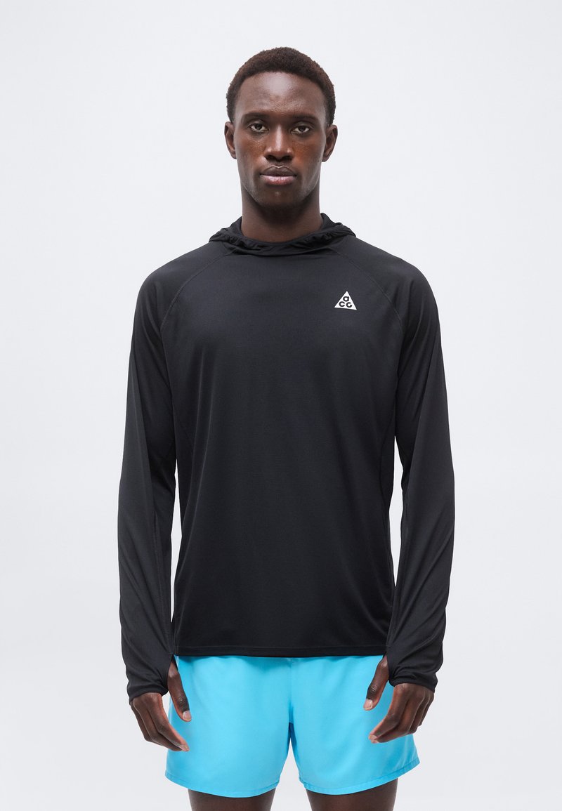 Άνδρας που φοράει μαύρο φούτερ με κουκούλα και λευκό τριγωνικό λογότυπο και φωτεινά μπλε σορτς, στέκεται μπροστά σε απλό ανοιχτό φόντο.