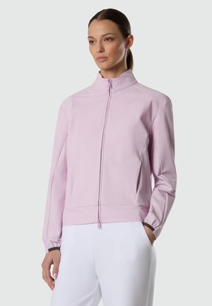 Donna con capelli castani raccolti in una coda bassa, indossa una giacca rosa chiaro con cerniera e pantaloni bianchi, in piedi con una mano in tasca.