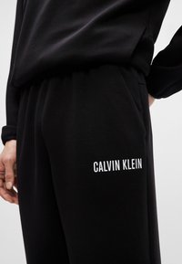Czarne dresy wykonane z miękkiego materiału z elastycznym pasem, z białym logo "CALVIN KLEIN" nadrukowanym na lewej nodze.