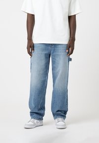 Jean en denim bleu clair avec une coupe ample, présentant un lavage délavé, des coutures contrastées et des poches latérales. Associé à des baskets blanches.