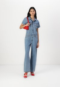 Denim overal s krátkými rukávy, dvěma kapsami na prsou a širokým střihem nohavic. V kombinaci s červenou taškou a červenými vysokými podpatky.