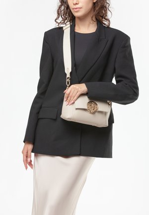 Donna con blazer nero e gonna crema tiene una borsa a tracolla in pelle color crema con fibbia circolare dorata.
