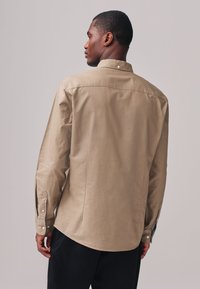 Beige overhemd met lange mouwen en een button-down kraag, afgewerkt met manchetten en een subtiele glans. Heeft een rechte achternaad en een getailleerde pasvorm.