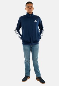 Marineblauwe sportjack met ritssluiting en witte driekleurige details op de mouwen, gecombineerd met lichtblauwe jeans en zwarte sneakers met bruine accenten.