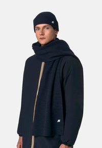 Cappello e sciarpa lavorati a maglia color blu navy scuro, con una texture a coste e un dettaglio con logo. Indossati con una giacca nera con cerniera, caratterizzata da una zip colorata.