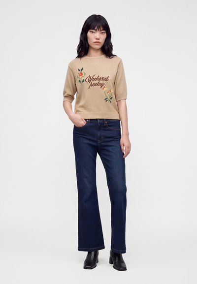 Femme aux cheveux noirs portant un pull beige avec broderie florale et texte, un jean droit bleu foncé et des bottines noires.