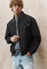 Mladý muž s kudrnatými vlasy, oblečený v černé bundě na zip přes šedé tričko a světle modré džíny, stojící před béžovým pozadím.