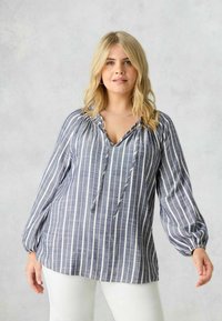 Blusa a maniche lunghe a righe in navy e bianco, con scollatura a V, dettagli con coulisse e polsini elasticizzati, abbinata a pantaloni di colore chiaro.