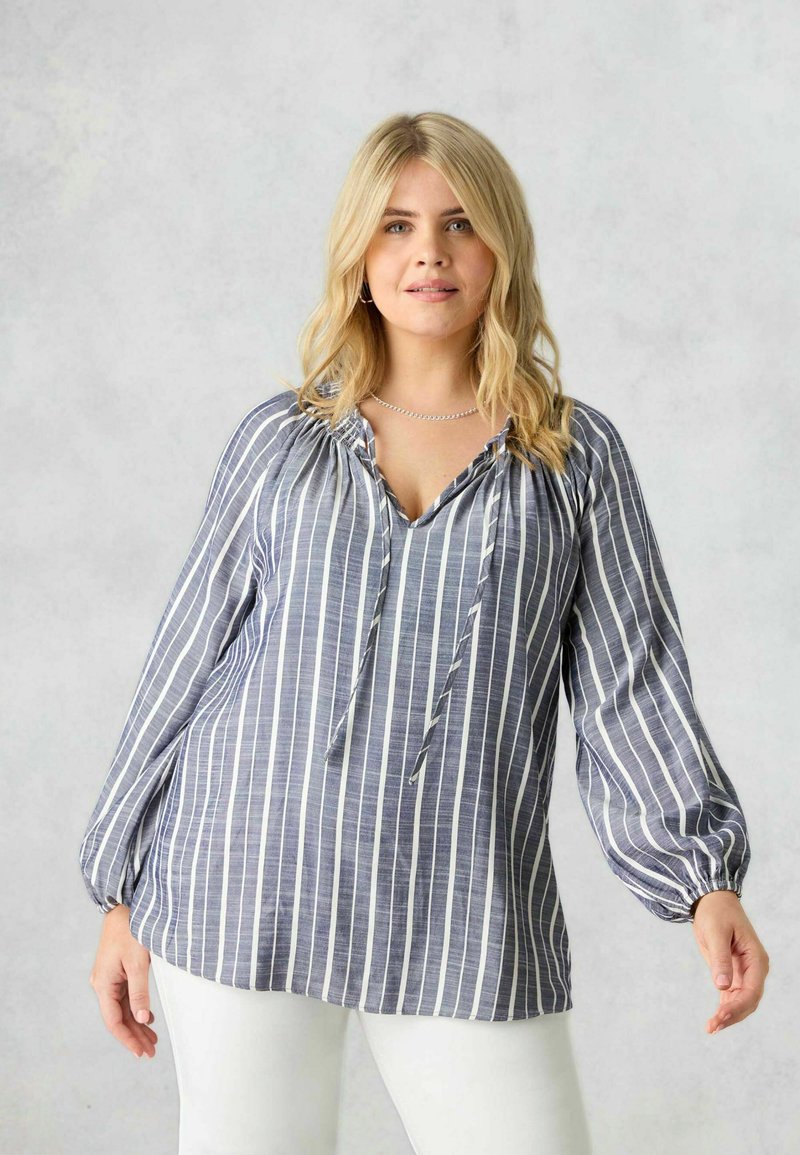 Blusa a maniche lunghe a righe in navy e bianco, con scollatura a V, dettagli con coulisse e polsini elasticizzati, abbinata a pantaloni di colore chiaro.