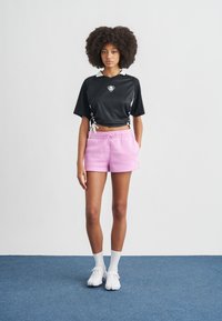 Μαύρο αθλητικό τοπ cropped με στρογγυλή λαιμόκοψη και λευκές λεπτομέρειες, συνδυασμένο με ροζ σορτς με κορδόνι. Το μοντέλο φοράει λευκά αθλητικά παπούτσια και κάλτσες.