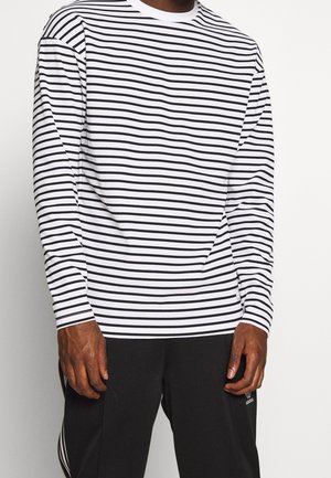 Mann trägt ein langärmeliges weißes Hemd mit schwarzen horizontalen Streifen und schwarze Adidas-Hosen mit weißen Streifen.