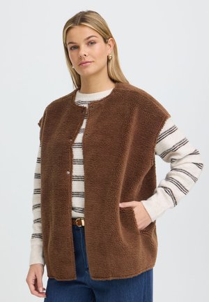 Kvinde iført en brun fleecevest over en hvid stribet langærmet sweater og blå jeans med den ene hånd i vestens lomme.