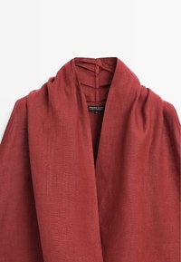 Veste en lin rouge rouille avec large col châle, montrant l'étiquette de la marque cousue à l'intérieur du col.