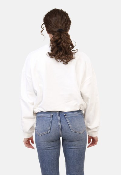 Felpa bianca over size con spalle scese indossata su jeans di denim blu a vita alta. Tessuto liscio con polsini e orlo a coste.