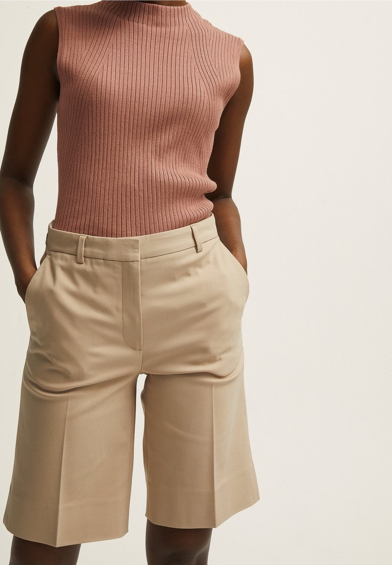 Hladký bezramenný top v jemné růžové barvě, doplněný o béžové šortky se zapracovanými kapsami a rovnými nohavicemi, s jemnou texturou.