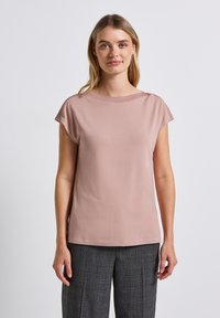 Kobieta o blond włosach, ubrana w fioletowawy top z krótkim rękawem i szare spodnie w kratę, stojąca na tle jednolitego białego tła.