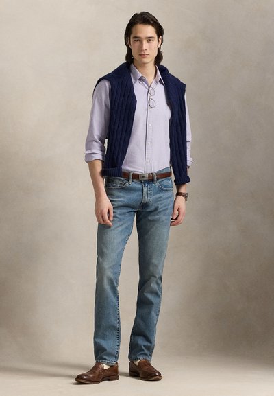 Γιλέκο σε σκούρο μπλε πλεκτό με κοχύλι πάνω από ένα ανοιχτό μοβ πουκάμισο με κουμπιά, σετ με ανοιχτό μπλε τζιν παντελόνι και καφέ δερμάτινα παπούτσια, κρατώντας γυαλιά στο χέρι.