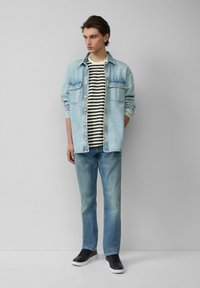 Lichtblauwe denim overhemd, gestreept zwart-wit t-shirt eronder, vervaagde blauwe jeans, zwarte sneakers met witte zolen, staande pose.