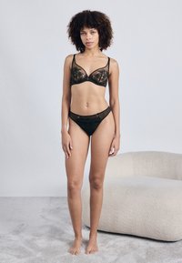 Μαύρο δαντελένιο bralette και αντίστοιχο μπικίνι με διακοσμητικά στοιχεία. Το bralette διαθέτει βαθύ ντεκολτέ και διαφανείς λεπτομέρειες από δίχτυ.