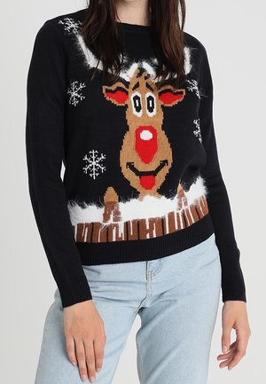 Femme portant un pull de Noël noir avec un renne souriant, des flocons de neige et une clôture enneigée, associée à un jean bleu clair.