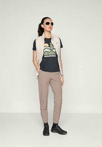 Lichtgewicht beige gewatteerde vest over een donker t-shirt met een natuurprint. Tan broek en zwarte schoenen maken de outfit compleet. Zonnebril inbegrepen.