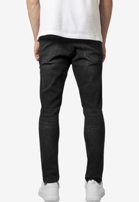 Černé džíny slim fit s hladkou texturou, vybavené dvěma zadními kapsami a zúženým střihem, kombinované s bílými teniskami.