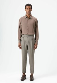 Lichtbruine lange mouwen blouse met knopen gecombineerd met lichtgrijze geplooide broek en donkerbruine loafers, alles op een neutrale achtergrond.