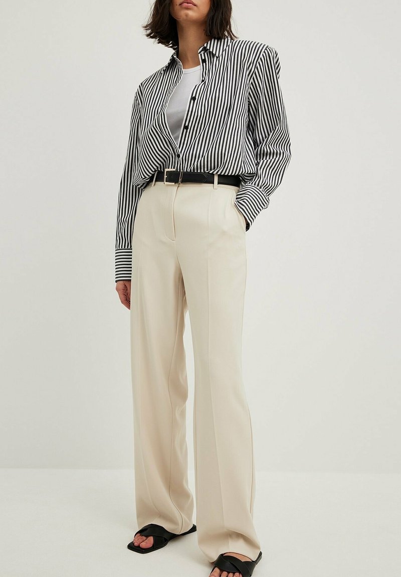 Chemise rayée noir et blanc, pantalon beige taille haute, ceinture noire et sandales à larges brides. Texture de tissu lisse, coupe décontractée.