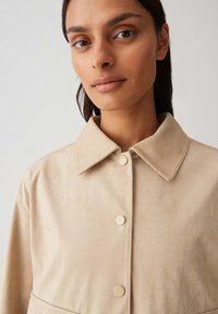 Giacca in suede beige con colletto classico, chiusura frontale a bottoni e texture liscia, che evidenzia la semplicità del design.