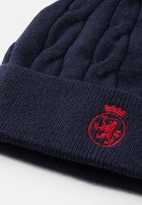 Granatowa dziana czapka z wzorem w warkocze, z prążkowanym mankietem oraz czerwoną haftowaną godłem z detalem lwa i korony.