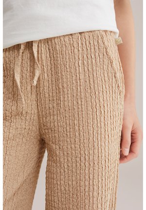 Beige textuurbroek met koordtouw in de taille, gedragen met een wit topje, waarbij de rechterzijde van de taille en hand zichtbaar is.