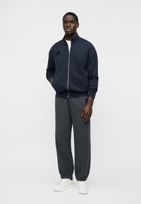 Homme portant une veste zippée bleu marine, un pantalon de jogging gris foncé et des baskets blanches, debout avec une main dans la poche sur un fond blanc.