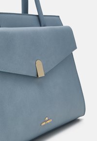 Sininen synteettisestä nahasta valmistettu käsilaukku, jossa on rakenteellinen muotoilu, läppäklipsi kultaisilla yksityiskohdilla ja brändäys etupuolella.