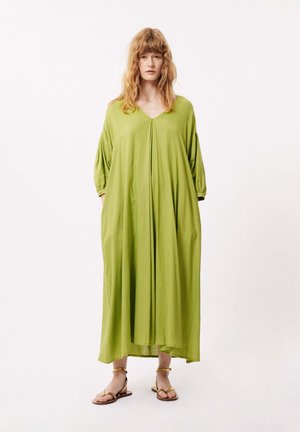 Robe verte longue et ample avec un décolleté en V, des manches ballon et des plis verticaux, fabriquée dans un tissu léger. Associée à de simples sandales marron.