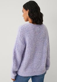 Vrouw met schouderlang donker haar, draagt zilveren oorbellen en een lavendelkleurige fluffy trui, gezien van achteren tegen een effen achtergrond.