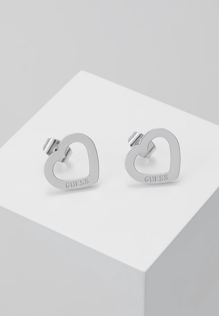 Boucles d’oreilles clous en forme de cœur en métal argenté avec ""GUESS"" gravé sur chacune, présentées sur une surface blanche inclinée.