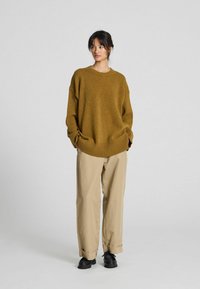 Overdimensioneret sennepsgul sweater med ribbet kant og manchetter, parret med lysebeige bukser og sorte sko, mod en ensfarvet baggrund.