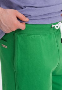 Pantalons de survêtement en coton vert avec taille à cordon, poches latérales et poignets côtelés, présentant une texture lisse et un minimum de détails de marque.