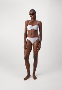 Hvit bikini med røde bølgekantdetaljer, med en stroppeløs topp og høylivs underdel, iført solbriller. Glatt tekstur, minimalistisk design.