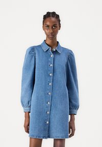 Jeansklänning med krage, knäppning fram och puffade långärmar. Tyget är ljust blått med en slät yta.