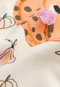 Tessuto morbido con un design a macchie arancioni e marroni, impreziosito da dettagli in pom-pom rosa, abbinato a illustrazioni di zucche su uno sfondo color crema.