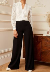 Bílá krajková blůza s výstřihem do V a dlouhými rukávy, v kombinaci s černými kalhotami s vysokým pasem a širokými nohavicemi a bílými teniskami. Hladká textura, elegantní design.