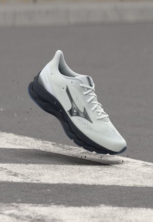 Balts sporta apavs ar melnu zolīti un Mizuno logotipu, gaisā virs reljefas ceļa virsmas ar baltām joslām.